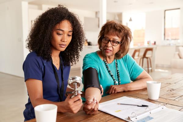 an older Black woman has her blood pressure taken in her home by a young Black woman health care professional